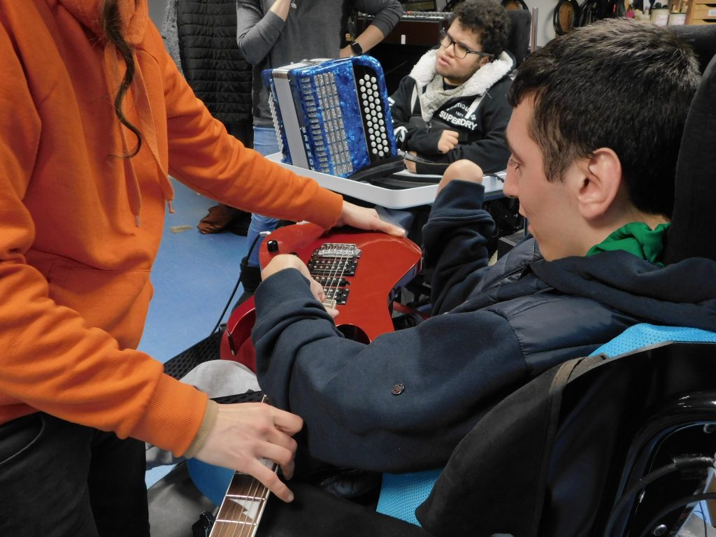 Clémentine montre au premier à un des participants comment jouer de la guitare électrique, et derrière on peut voir un autre participant qui s'essaie à l'accordéon